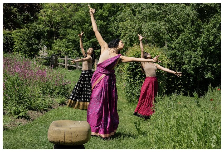 Three women are dancing in a rural location. They all wear brightly coloured costumes