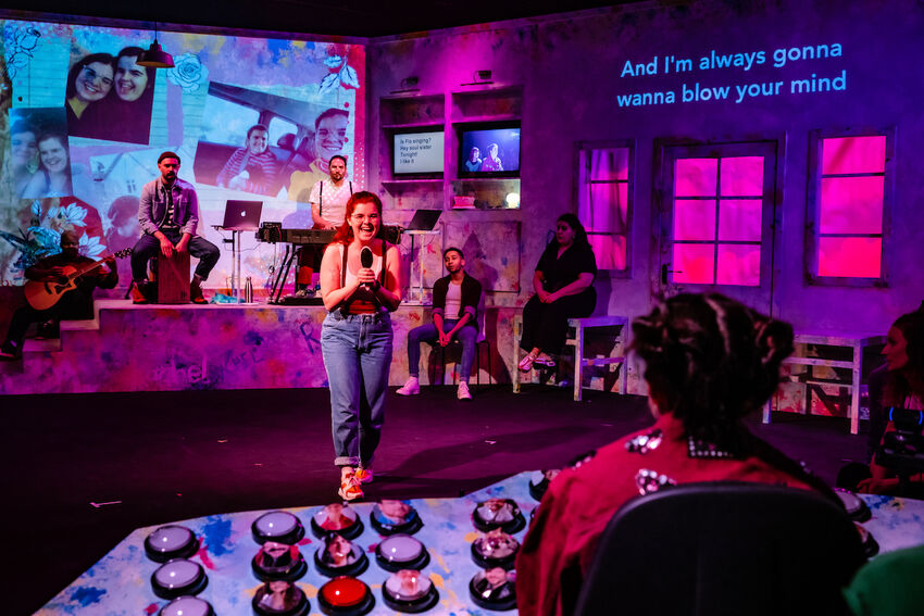 Looking over the shoulder of Rachel and a mixing desk, a person sings into a hairbrush with projections of a photo collage of them
