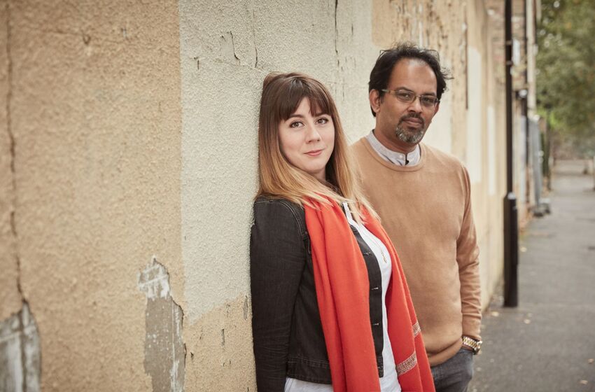 Helen Anahita Wilson and Shahbaz Hussain pose on the street against a beige crumbling wall
