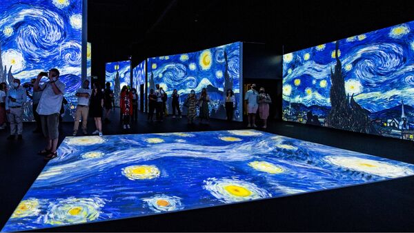 A group of audience members stand around an interpretation of Van Gogh's The Starry Night. It is projected onto the floor and screens which surround them 