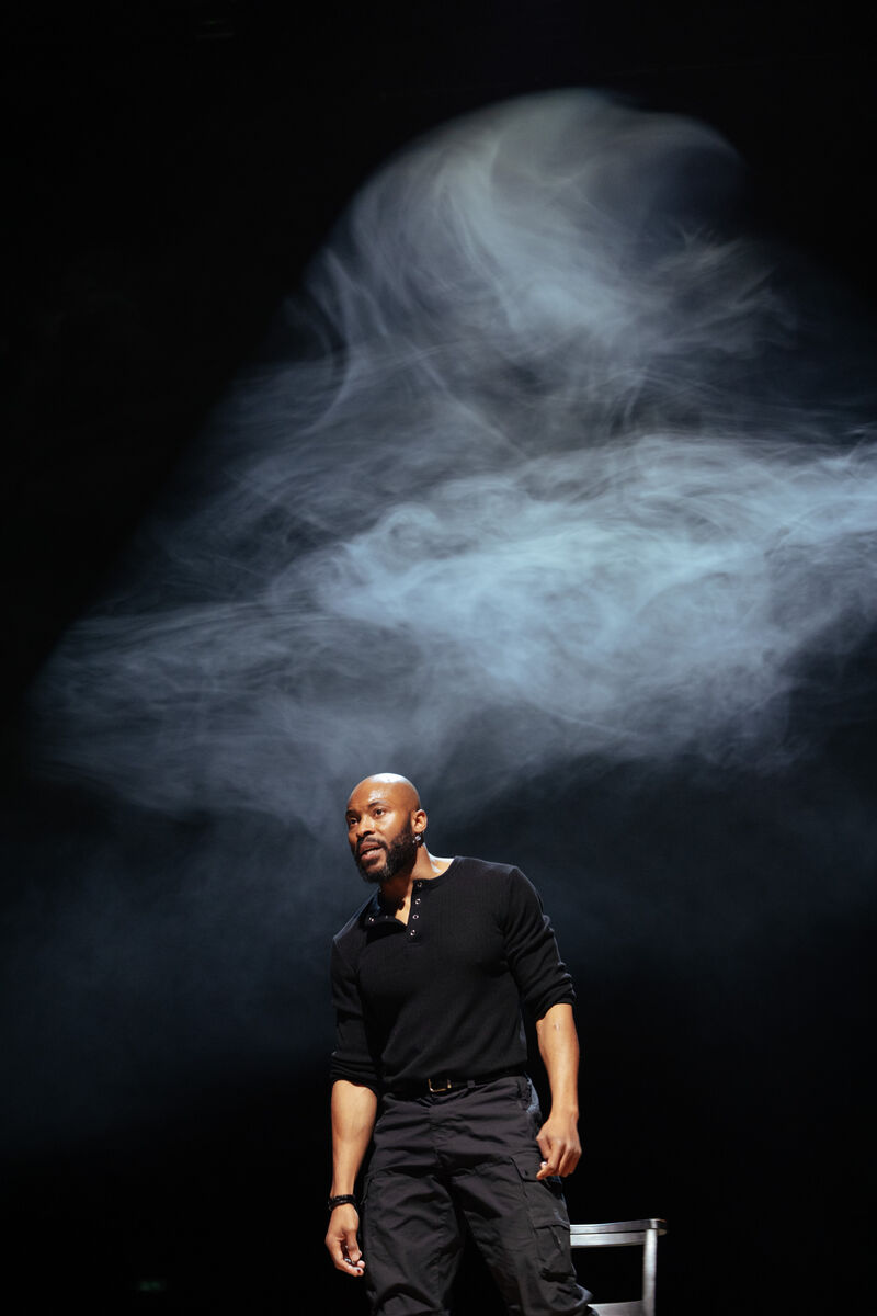 Man standing on stage in black shirt and trousers. He is surrounded by haze