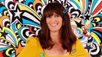 Liz Pichon smiles at camera. She is wearing a bright yellow dress, has a colourful clip in her hair and is standing in front of brightly patterned wallpaper