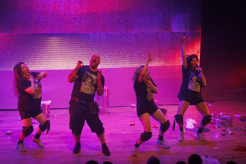 On stage picture of four performers singing and dancing in extravagant poses, the floor is littered with cans and trash