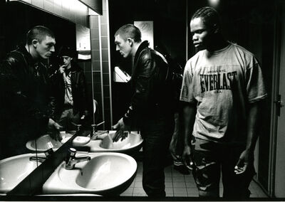 A black and white photo of a black man wearing a t shirt and a white man wearing a leather jacket standing in front of two bathroom sinks and looking at themselves in the mirror