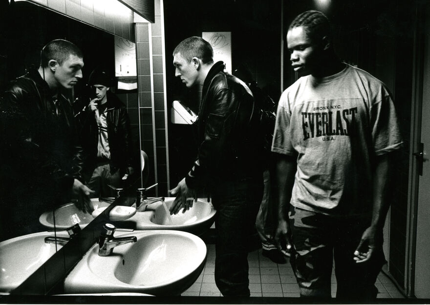 A black and white photo of a black man wearing a t shirt and a white man wearing a leather jacket standing in front of two bathroom sinks and looking at themselves in the mirror