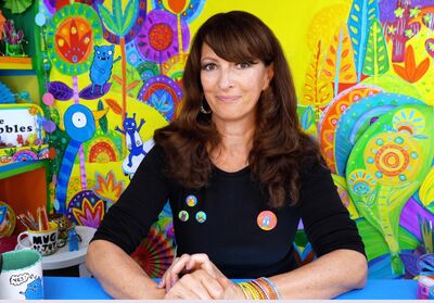 Liz Pichon sitting in front of a colourful background of her illustrations, she is smiling at the camera with her hands together on the table