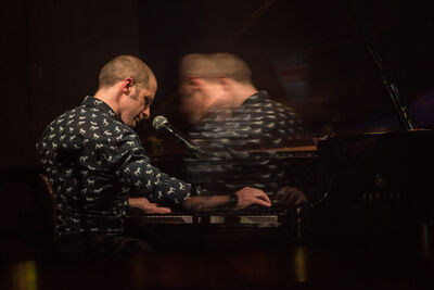 Ivo Neame, a white man with a buzz cut wearing a black shirt with white dogs on it, sits and plays the piano. An image effect has created a blurred mirror image to the right of the photo