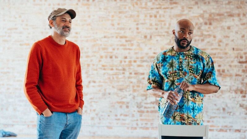 Omar Elerian, a theatre director in a red jumper, stands next to performer Arinze Kene who is holding a large bottle of water