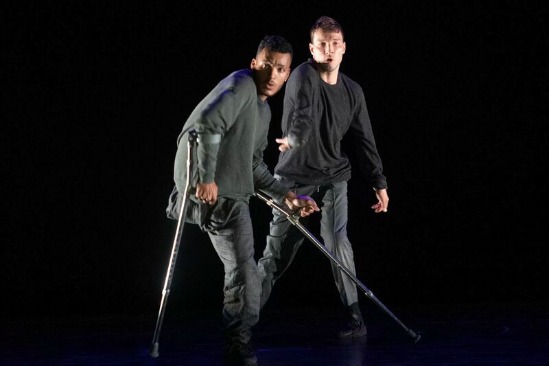 Two dancers wearing dark clothes dance on a stage. The one closest to the front is using crutches.