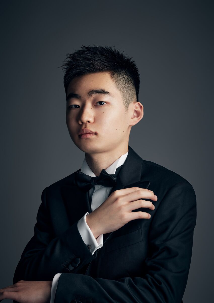 A young Japenese man with short black hair stands with his arm across his chest. He wears a black suit and black bow tie.