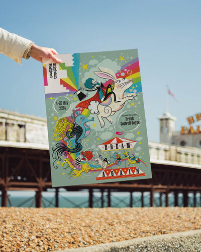 A hand holding up a print of the Brighton Festival 2024 brochure cover in front of Brighton Pier
