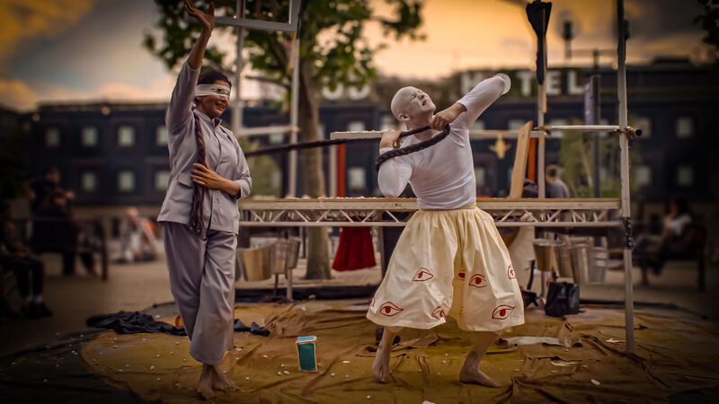 Two dancers on an outdoor, flat stage. One is all in white pulling rope with a strained face, the other blindfolded and in a light blue outfit. 