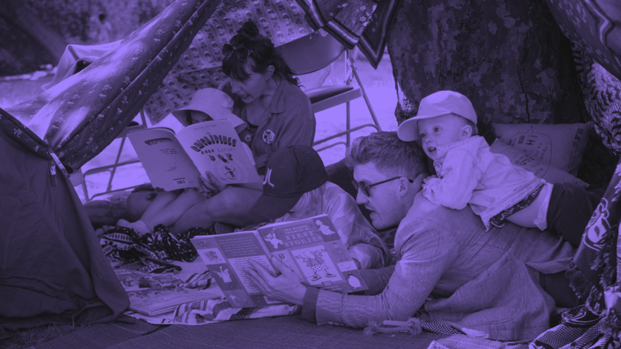 A man in sunglasses lay on his front reading a book to his child that lay on his back