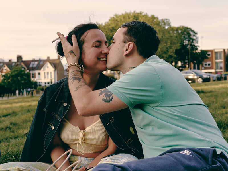 A woman smiling as a man kisses her while sitting in a park