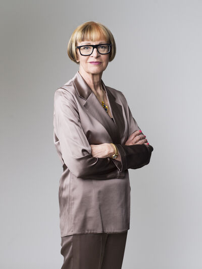 Philippa Perry stands smiling against a grey background, with her arms crossed