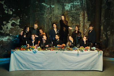 Twelve people sit on one side of a long table with a white tablecloth covered in food, reminiscent of the Last Supper