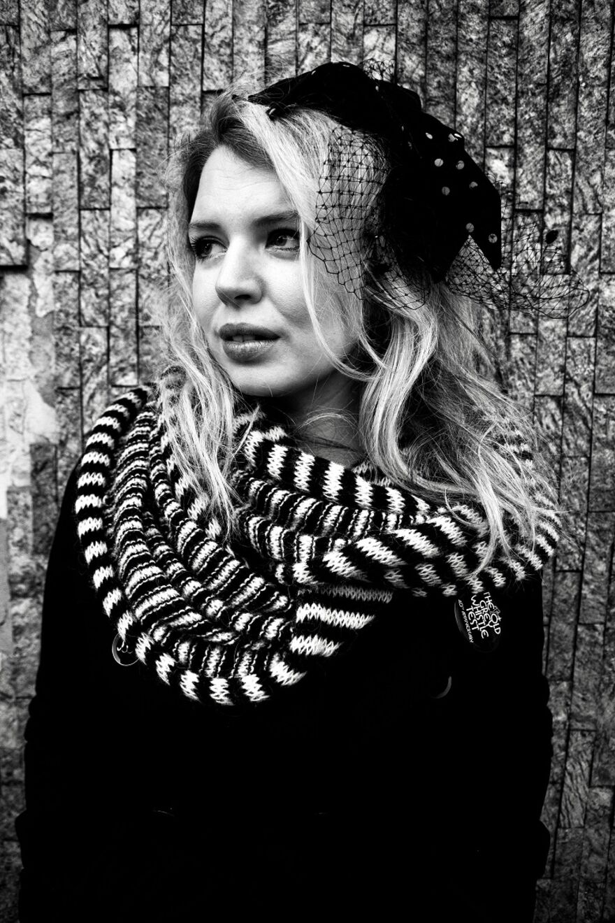 A white woman in a black fascinator style hat, black and white scarf and black top