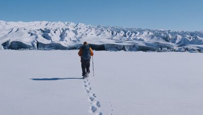 A singular person in a bright orange coat walks through a barren arctic landscape. They are leaving footprints in the snow and there are mountains in background