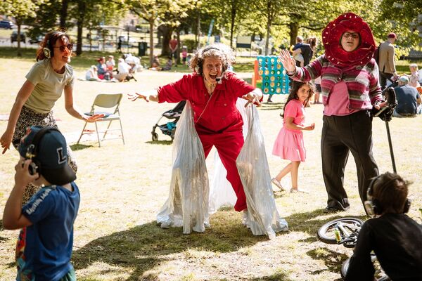 A person in a red boiler suit and silver wings stands in a field entertaining children. There are other children and adults around wearing headphones, and in the background are giant party games