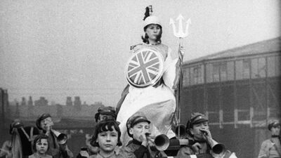 A woman dressed in armour, holding a trident and a shield with the United Kingdom flag on it. Below her are some young people, some of who are blowing in to horns.