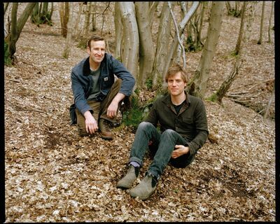 Two men sit in a wooded area. The ground is covered in leaves and they look towards the camera