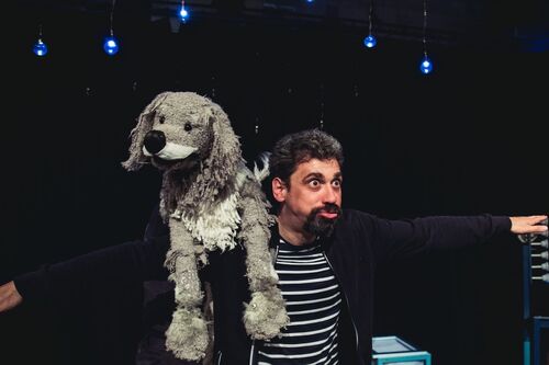 Darren East stands on stage. He has a puppet of a dog over his shoulder and has his arms out to the side like an airplane. He is pulling a funny face and wears a stripy black and white t-shirt
