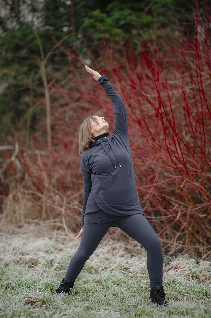 A woman is in a rural environment. She is in a yoga pose known as reverse warrior with her legs in a lunge position, one arm on her back leg and the other reaching into the air in a slight backbend