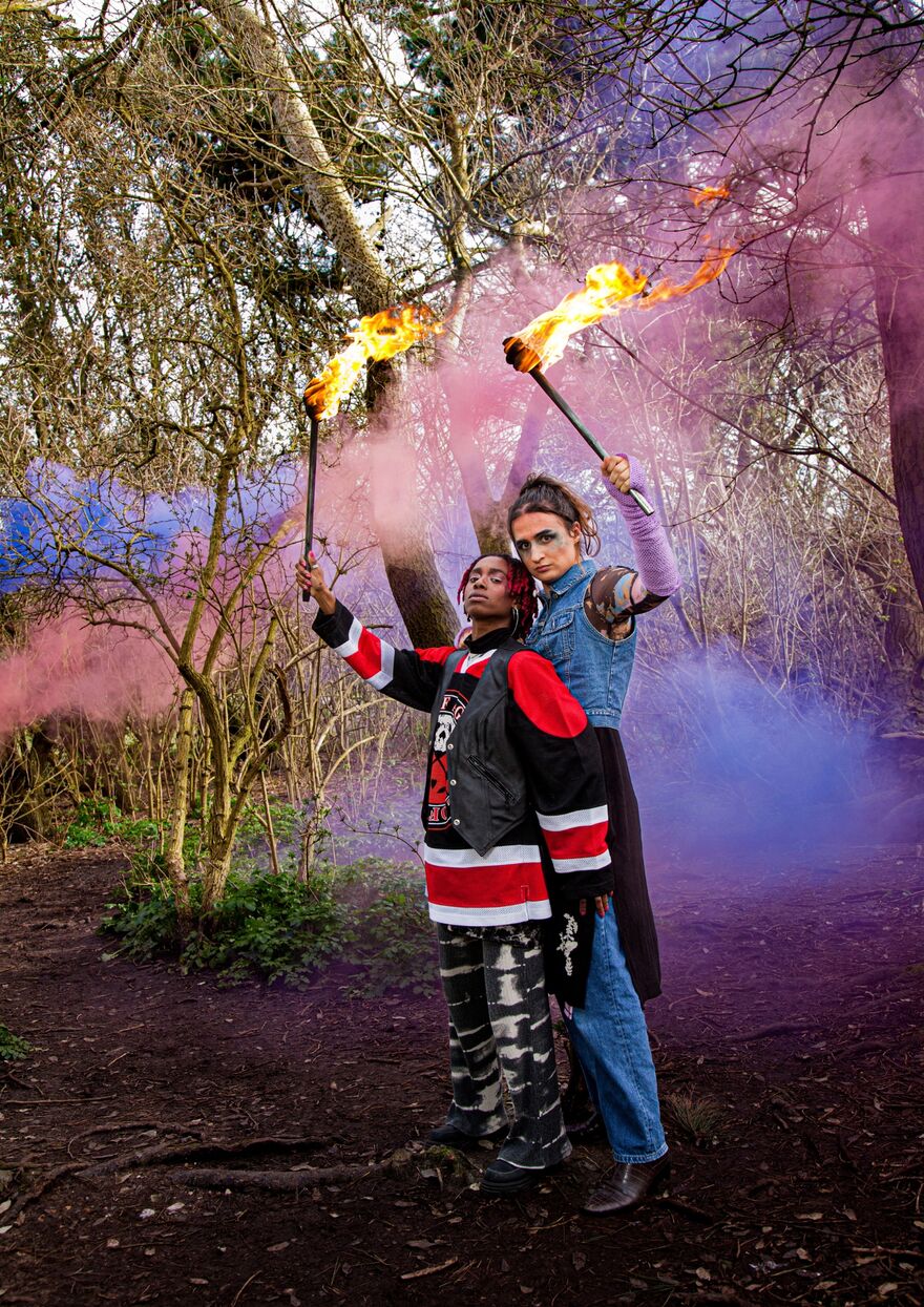 Two performers stand outside in the woods together closely, surrounded by pink and blue smoke, holding flaming torches  