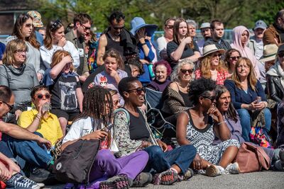 A wide shot of a mixed group of people sat and standing watching an outdoor theatre show