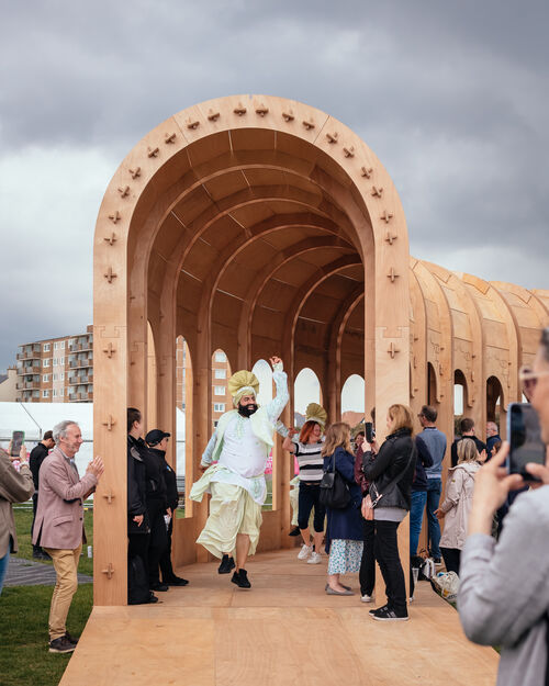 A Bhangra dancer wearing white and green Indian clothing leads a line of people through the Riwaq