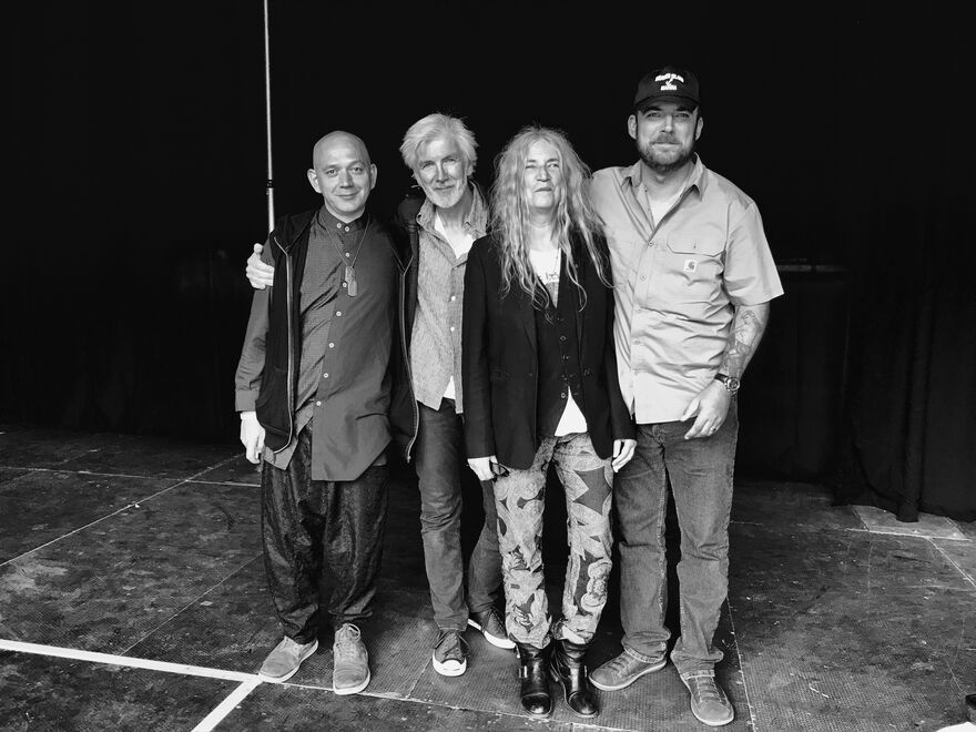 Black and white photo of the Patti Smith Quartet. Left to right there's a short bald man wearing baggy trousers and a long shirt, an older man with grey hair wearing a shirt and jeans, an older lady with long curly grey hair wearing patterened trousers and a black blazer, and a tall younger man with a beard wearing a baseball cap, shirt and casual trousers