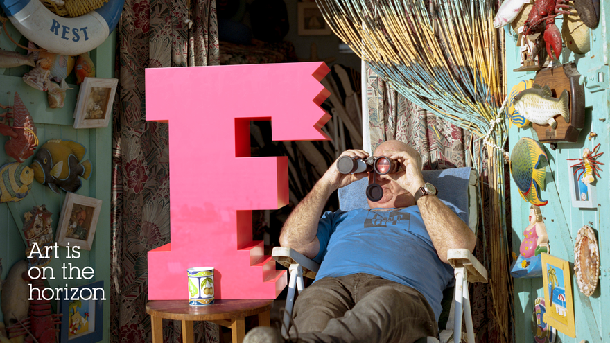 A person with binoculars sitting on a deck chair, with a giant pink 'F' next to them