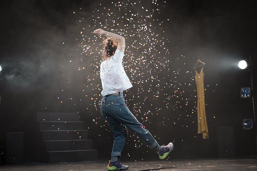 A woman in a white t-shirt and jeans stands with one leg slightly raised, and one arm raised. She is surrounded by confetti.