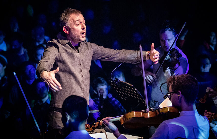 A person in a grey top, with short greyish hair conducts an orchestra