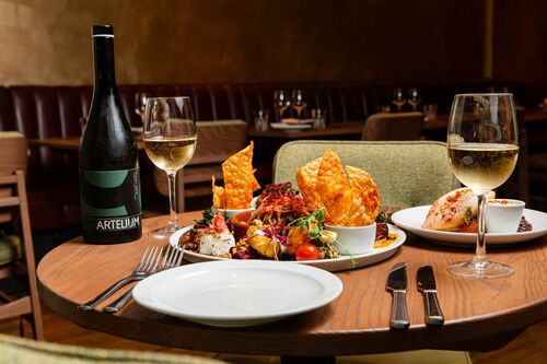 A wooden table filled with plates of delicious food, two glasses of wine and a bottle of wine