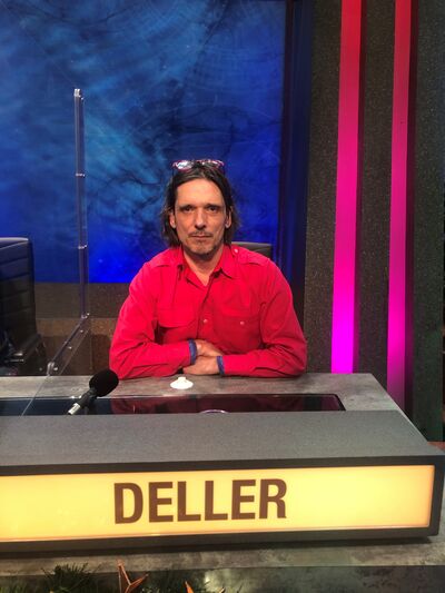 A white man with mid-length hair sits at a desk, in front of him is a sign that reads 'DELLER'