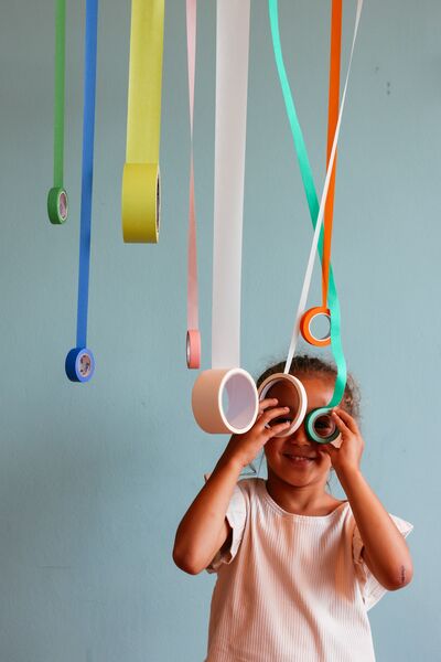 Roles of tape hang down from above the frame. A child looks through two of the tape roles like glasses, smiling