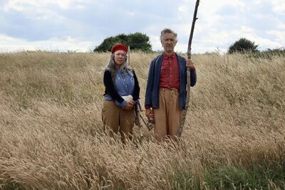 A man and a woman stand in a field of tall grass, they have plain, staring expressions. The man is holding a large stick, and the woman is holding a large stone.
