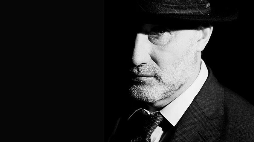 Black and white image of Jah Wobble wearing a suit and a brimmed hat - a shadow covers half his face