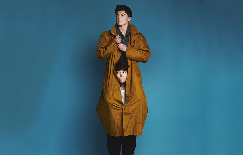 Two young looking queers, one standing on the others shoulders, whilst they wear a long brown coat