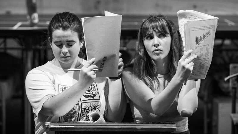 Two girls sitting at a desk holding open paper booklets up over the side of their faces