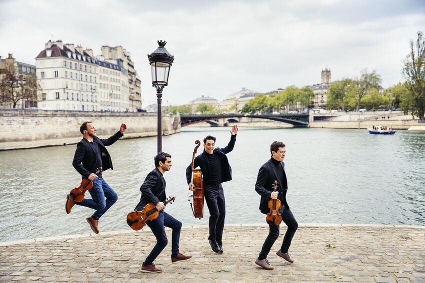 Four men in suits jump by a river holding stringed instruments