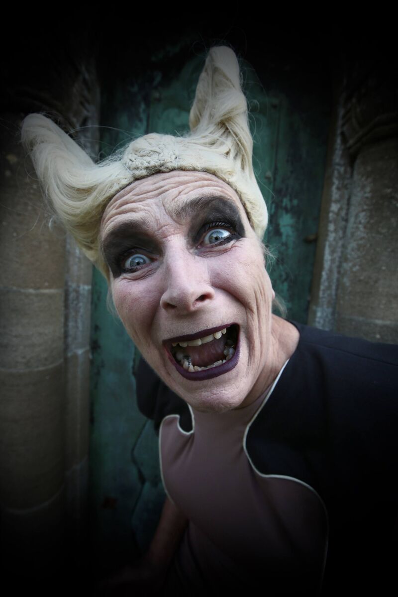 A close-up of Liz Aggiss screaming towards the camera, a blonde horned wig, intense black eyeshadow and deep purple lipstick