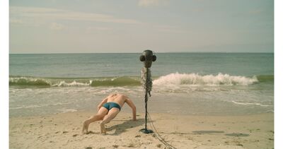 A still from 32 Sounds: A sand beach with a speaker on a stand, a man does a press-up beside it