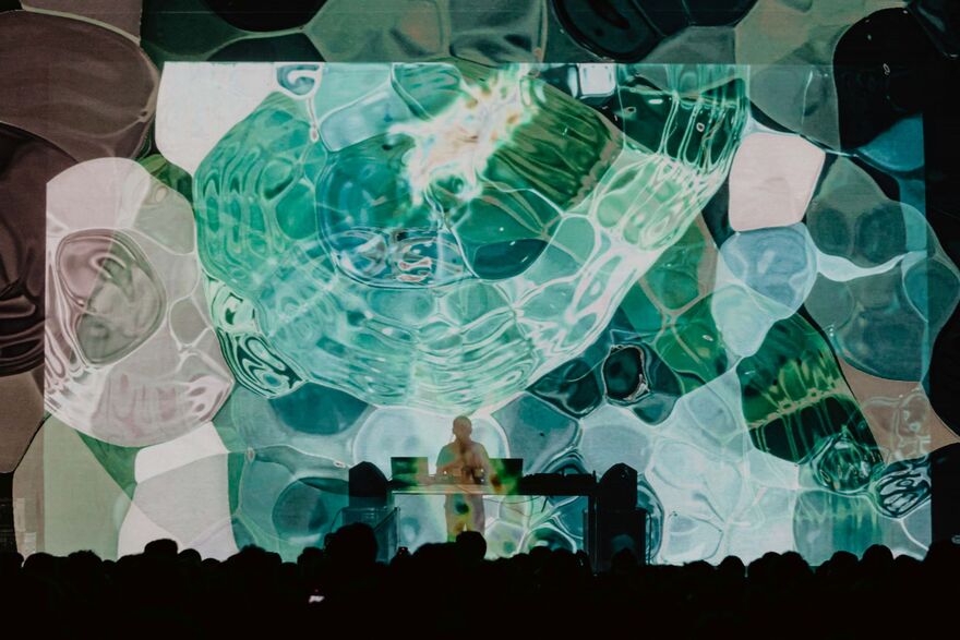 Max Cooper at a DJ deck, silhouetted with a screen of projections with blue, green and white blobs