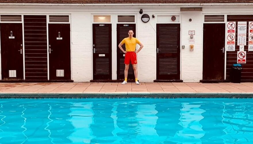 A man stands at the centre of a poolside wearing a yellow top and red shorts, in front of blue water