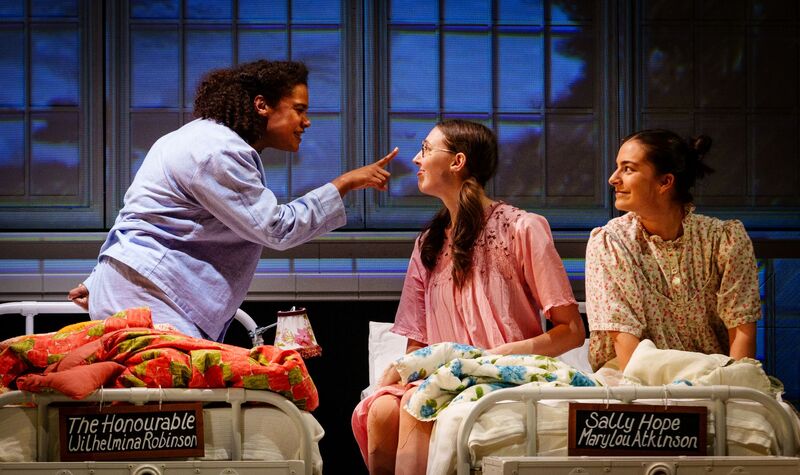 Three people sitting on boarding school beds. One is holding their finger up to another in an angry manner