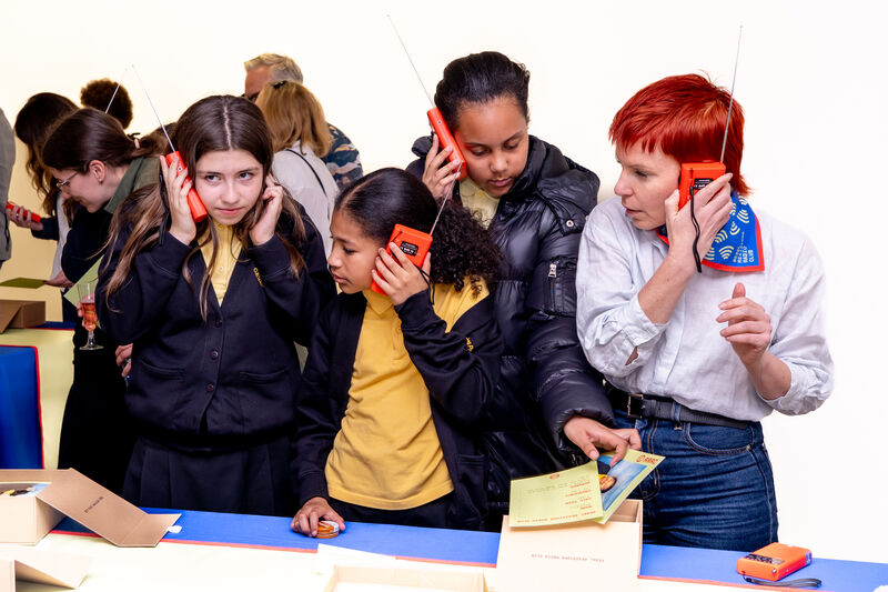 Mulitple young girls stand around a table listening to orange radios