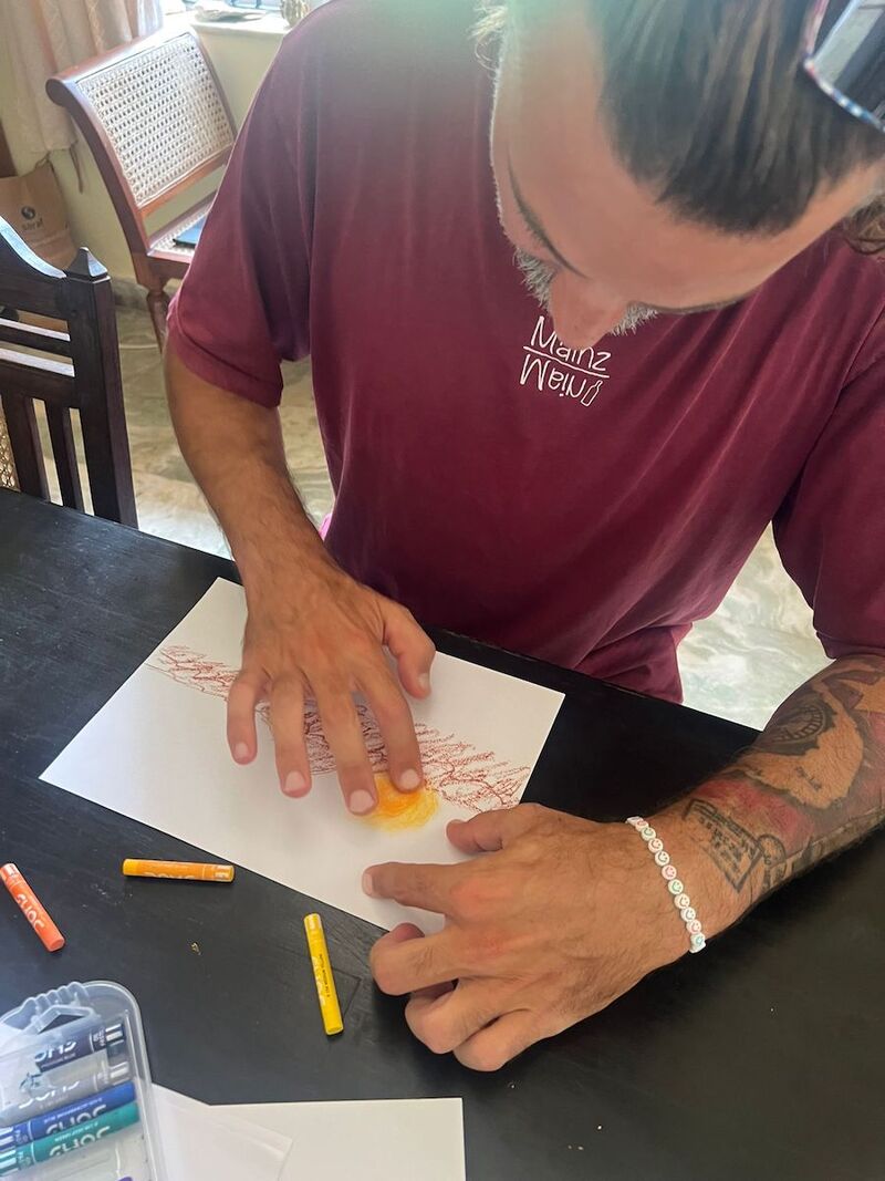 An older man sits at a table using yellow oil pastels to draw a sun. He is smudging the oil pastels with his finger