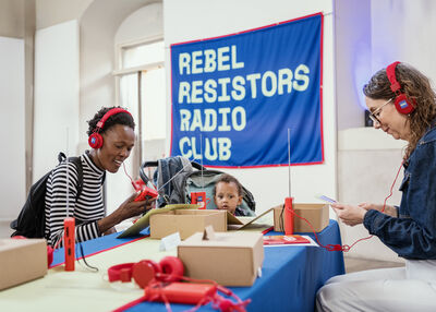 Two adults and a child sit around a table as they make their own radio, a banner in the background reads Rebel Resistors Radio Club